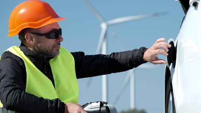 Windmills generate alternative energy for ev. Engineer in orange helmet opens cap and connects plug to electric car socket for charging on foreground