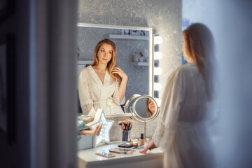 A young, slender blonde woman looks in the mirror in the bedroom. She is wearing a beautiful silk white robe.
