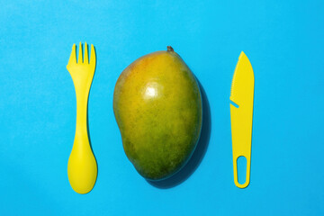 Whole mango fruit with a plastic knife and fork on a blue background. View from above