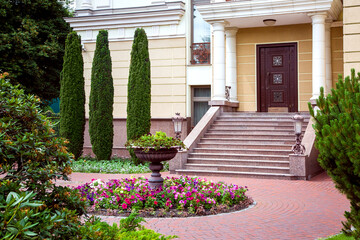 Fototapeta na wymiar granite staircase with iron retro street lighting lanterns on threshold with columns at entrance to door of house on backyard with landscape design and stone flowerpot, exterior of facade with thuja.