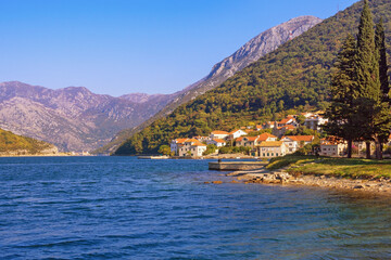 Fototapeta na wymiar Sunny autumn day, beautiful Mediterranean landscape with small seaside village against mountains. Montenegro, Adriatic Sea. View of Kotor Bay and Lepetane village