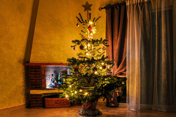 A Christmas tree decorated with toys and lights stands in the semi-darkness of an antique room with uneven beige walls with curtains and a brick fireplace in the background.