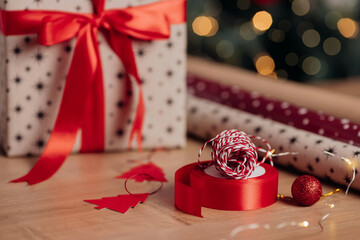 Beautiful xmas gift box wrapped in kraft paper and red ribbon standing on background of christmas tree and lights. Presents preparation in winter holidays