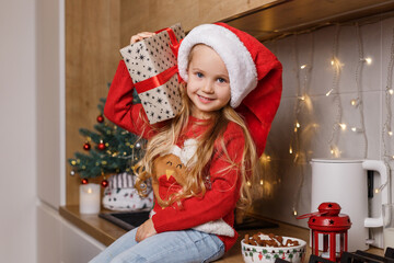 Happy little girl smiling and holding in hands wrapped gift box with bow on the background of christmas tree. Funny kid in red santa's hat having fun on winter holidays at cozy decorated home kitchen