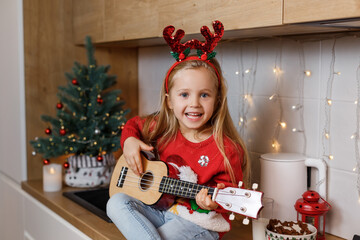 Happy little girl in red horns playing ukulele on the background of christmas tree. Talent kid having fun on winter holidays at cozy decorated home kitchen