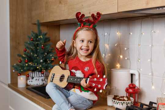 Happy little girl in red horns playing ukulele on the background of christmas tree. Talent kid having fun on winter holidays at cozy decorated home kitchen