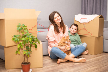 A young beautiful mother with her son moved to a new apartment and are sitting against the background of boxes with their fat ginger cat.