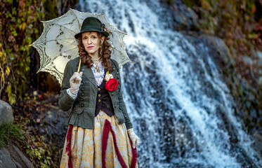 Woman in a 19th century suit with an umbrella against the background of a waterfall in autumn