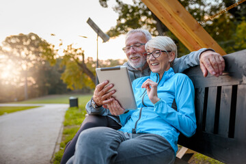 Smiling senior active couple sitting on the bench in the park looking at tablet computer. Using...