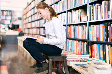 Preteen girl sitting on small wooden step ladder browsing textbook in library
