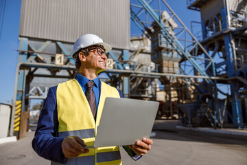 Joyful matured man in work vest holding laptop and smiling while standing on territory of production plant