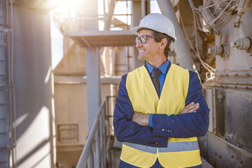 Joyful matured man wearing safety helmet and work vest while keeping arms crossed and smiling