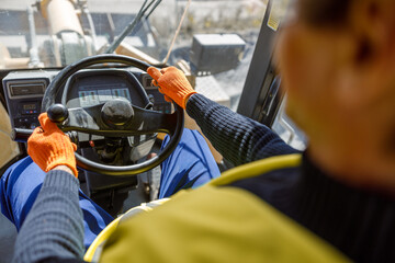 Close up of man in work gloves sitting in vehicle driver cabin and placing hands on steering wheel