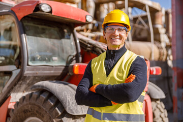 Cheerful matured man in safety helmet and work vest looking at camera and smiling while keeping...