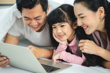 Cheerful young Asian family of handsome father and beautiful mother lying on bed with cute little daughter laughing while watching a comedy movie on laptop and relaxing at home