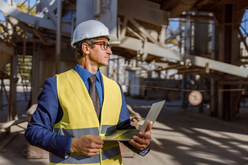 Matured man engineer wearing safety helmet and work vest while holding modern notebook