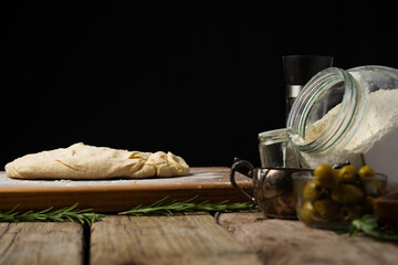 On a cutting board, kneaded dough, ingredients for making focaccia, pie, pizza nearby. Black background, rough wood texture. There are no people in the photo. There is an empty space for insertion.