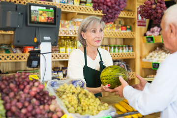 Senior saleswoman standing at counter in greengrocer and serving man customer who purchasing fresh melon.