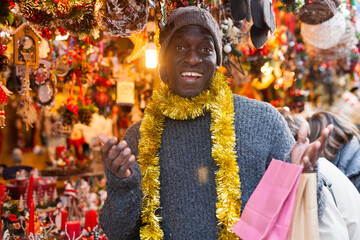 Portrait of laughing African-American guy holding bags after shopping on outdoor Christmas market.