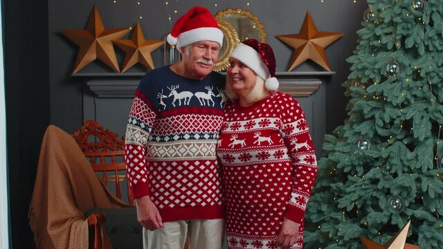 Portrait of happy senior couple in Santa Claus hats celebrating Christmas in cozy living room at home, looking into camera and hugging. Elderly people grandparents celebrate Christmas. Holiday Mood