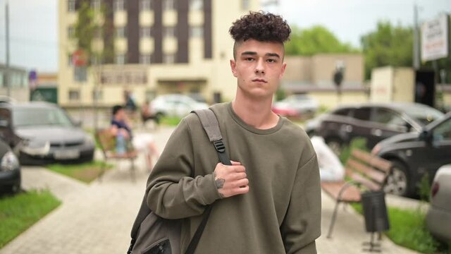 A young man with a backpack walks along the city alley and smokes an electronic cigarette. Slow motion. Modern stylish guy walking around the city