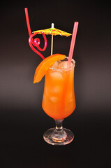 Tall glass of mango cocktail with ice, tropical fruit slice, straws and umbrella on black background.