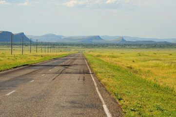 Straight two-lane asphalt road going through the endless steppe along the power line to meet the mountain range.