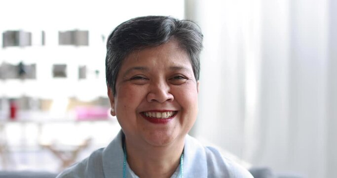 Optimistic mature Asian woman with short hair looking at camera and laughing at joke in sunlit room