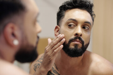 Serious bearded man looking at mirror when applying lotion or foundation in the morning