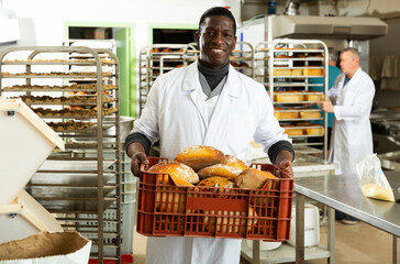 Skillful African American baker working in small bakery, carrying fresh baked bread in box ..