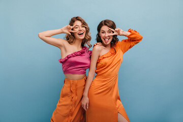 Two funny caucasian young girlfriends having fun and showing common gesture meaning victory or peace. On blue background funny girls show their tongues are dressed in bright clothes.