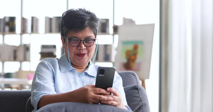 Happy mature Asian woman in glasses browsing social media on cellphone and laughing at joke while relaxing on couch in sunlit living room in weekend at home