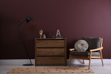 Elegant room interior with stylish chest of drawers, floor lamp and comfortable armchair near brown wall