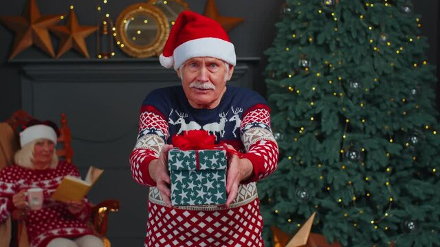 Elderly grandfather old man wears red Christmas sweater presenting Christmas gift box stretches out hands in cozy living room at home. Happy old mature family New Year Xmas celebration holiday eve