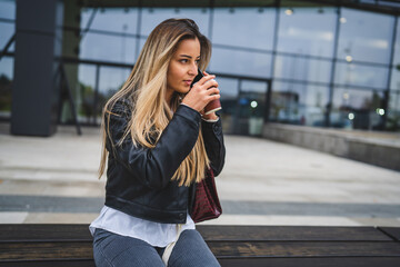 one young caucasian woman female student sitting in front of the university in autumn day holding a cup of coffee waiting while looking to the side holding mobile phone talking real people copy space