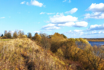 Beautiful autumn landscape in clear, sunny weather. Yellowed grass and trees on the steep bank of a blue river under a bright, blue sky with clouds.

