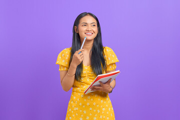 Smiling young Asian woman holding pen and notebook, looking up isolated on purple background