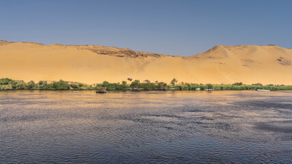 The Nile River flows calmly. Ripples on the water. There is green vegetation on the shore. High sand dunes against the blue sky. Egypt. Aswan