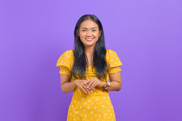 Smiling young Asian woman keeps hand together and feels optimistic on purple background