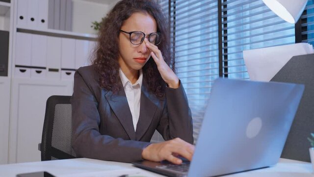 Exhausted Latino business woman hurt eye while using laptop computer. 