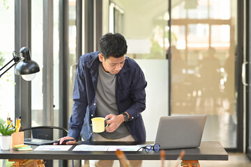 Young asian man graphic designer standing at office desk and using laptop computer.