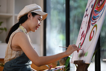 Happy young asian woman painting picture on canvas with oil paints in her studio.