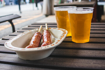 Yummy sausages on skewers and two cups of cold beer are on the wooden table.