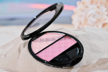 Fototapeta na wymiar open face blush in pink shades and mini brush. decorative cosmetic on sandy beach next to shell. make up blusher for summer.