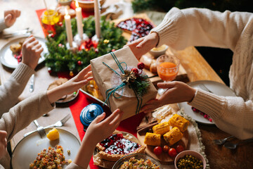 Close-up top view of unrecognizable loving parent giving festive box with Christmas present to happy son sitting at dinner feast table during holiday family party, selective focus, blurred background