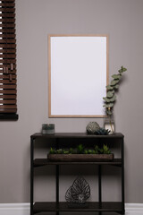 Empty frame hanging on grey wall over wooden console table with decor. Mockup for design