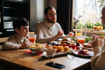 Side view of happy young family enjoying at festive Christmas table during holiday family party. Cheerful mother, father and little son having dinner xmas party, eating and chatting at home.