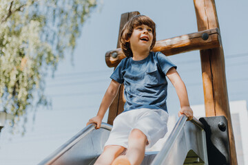 cheerful little boy in a blue T-shirt and white shorts rolls down a metal slide in city park. child walks outdoors. healthy active recreation. lifestyle. space for text. High quality photo