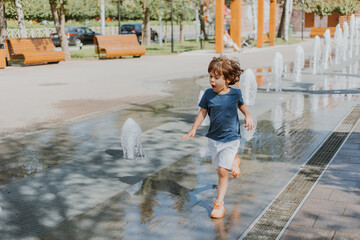 little boy is running in the street and playing with the water jets of a fountain spouting from the...