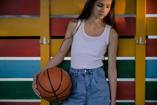 beautiful small young girl in white tank top and denim blue jeans retro fashion holding basketball ball on color wooden cloakroom background during summer leisure recreation sport activity